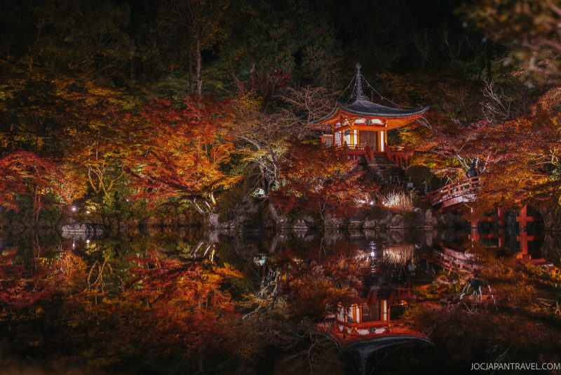 A brightly lit Japanese temple, reminiscent of ancient pagodas, is surrounded by autumn trees and reflected perfectly in a still pond at night. A curved bridge leads to the temple, while colorful foliage creates a serene, picturesque scene echoing the tranquil beauty of Kyoto Daigoji.