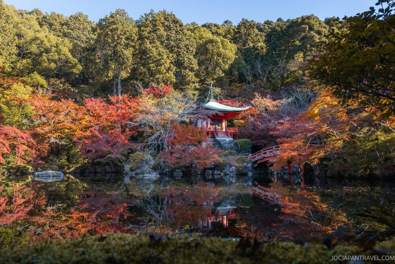 A small red pagoda and arched bridge surrounded by autumn trees are reflected in a calm pond, with a forested background under clear skies, reminiscent of the serene beauty found at Kyoto Daigoji, where ancient pagodas blend harmoniously into the natural landscape.