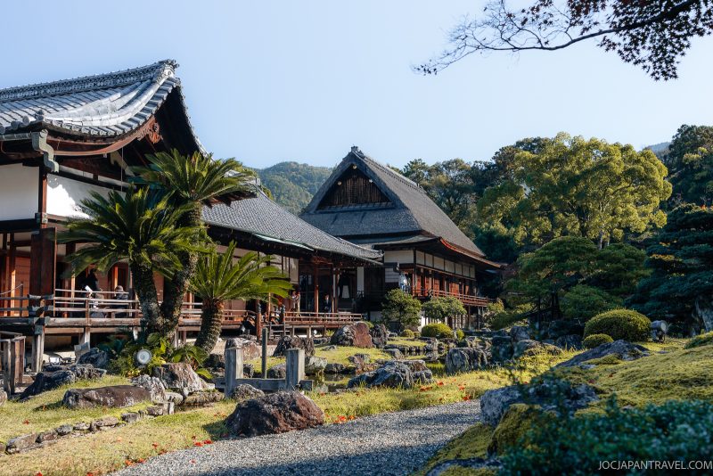 Traditional Japanese wooden buildings with tiled roofs surrounded by manicured gardens, trees, and stone paths under a clear blue sky, set against a backdrop of green hills and ancient pagodas reminiscent of Kyoto Daigoji—an inviting scene for those who enjoy tranquil strolls or mountain hikes.