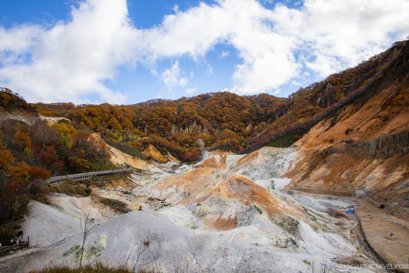 A valley with white and orange rocky terrain, surrounded by autumn trees under a partly cloudy sky. A wooden walkway curves along the left side of the scene.