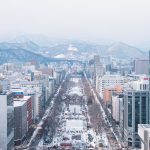 The Ultimate Hokkaido Itinerary: 5, 7, and 12-Day Routes A snowy cityscape with tall buildings on both sides of a wide, tree-lined avenue, leading towards distant snow-covered mountains under a cloudy sky.