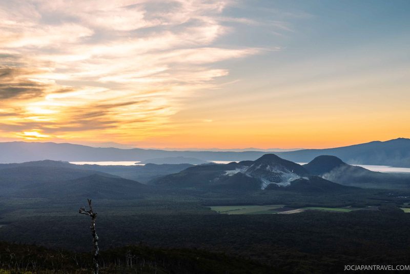 Sunset over a mountainous landscape with rolling hills, a glowing sky, and soft clouds. A single bare tree stands in the foreground, and distant lakes or mist appear on the horizon.