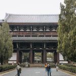 Large traditional Japanese temple gate with dark wooden beams, white walls, and a tiled roof, framed by tall green trees. Set within the historic grounds of Tofukuji, a renowned Kyoto Zen Temple, several people walk and stand in front of the gate on a gravel pathway.