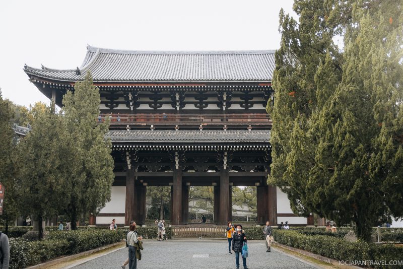 Large traditional Japanese temple gate with dark wooden beams, white walls, and a tiled roof, framed by tall green trees. Set within the historic grounds of Tofukuji, a renowned Kyoto Zen Temple, several people walk and stand in front of the gate on a gravel pathway.