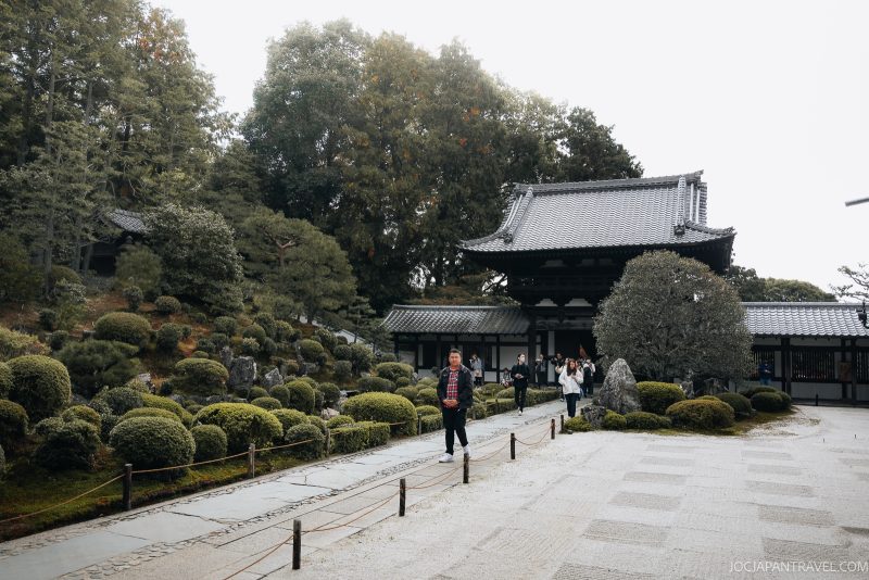 People walk on a stone path past carefully manicured shrubs and trees toward Tofukuji, a traditional Zen Temple in Kyoto with a tiled roof, surrounded by lush greenery.