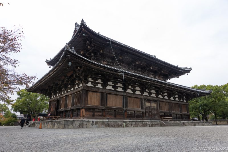 A large, traditional wooden Japanese temple with ornate roof details sits on a stone foundation, surrounded by trees and a gravel courtyard under a cloudy sky. Inspired by the architecture of Toji Temple in Japan, which is renowned for having the tallest wooden pagoda in the country, the scene captures a timeless sense of tranquility and grandeur.