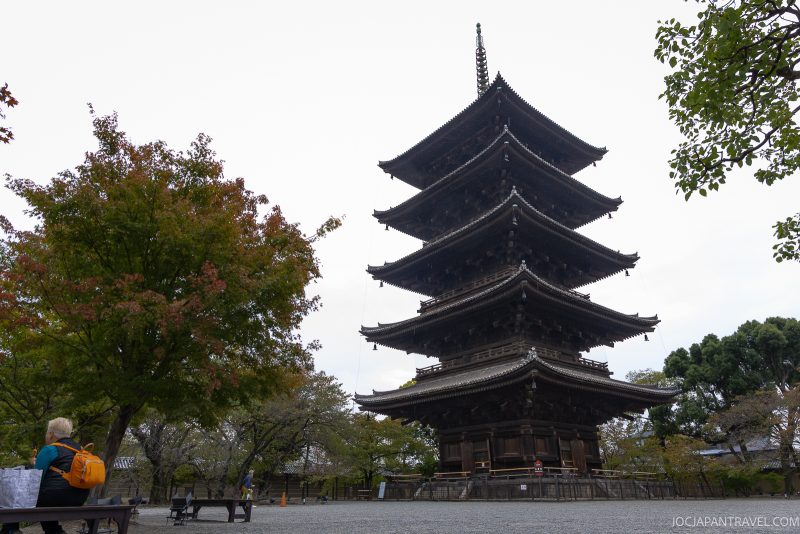 A five-story traditional pagoda, known as Japan’s Tallest Wooden Pagoda at Toji Temple, stands tall among trees, with a person sitting on a bench in the foreground and cloudy skies above.