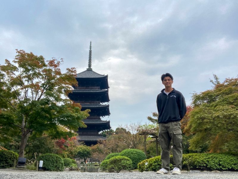 A person stands in a garden with trees and shrubs, with Japan’s Tallest Wooden Pagoda at Toji Temple rising in the background under a cloudy sky.
