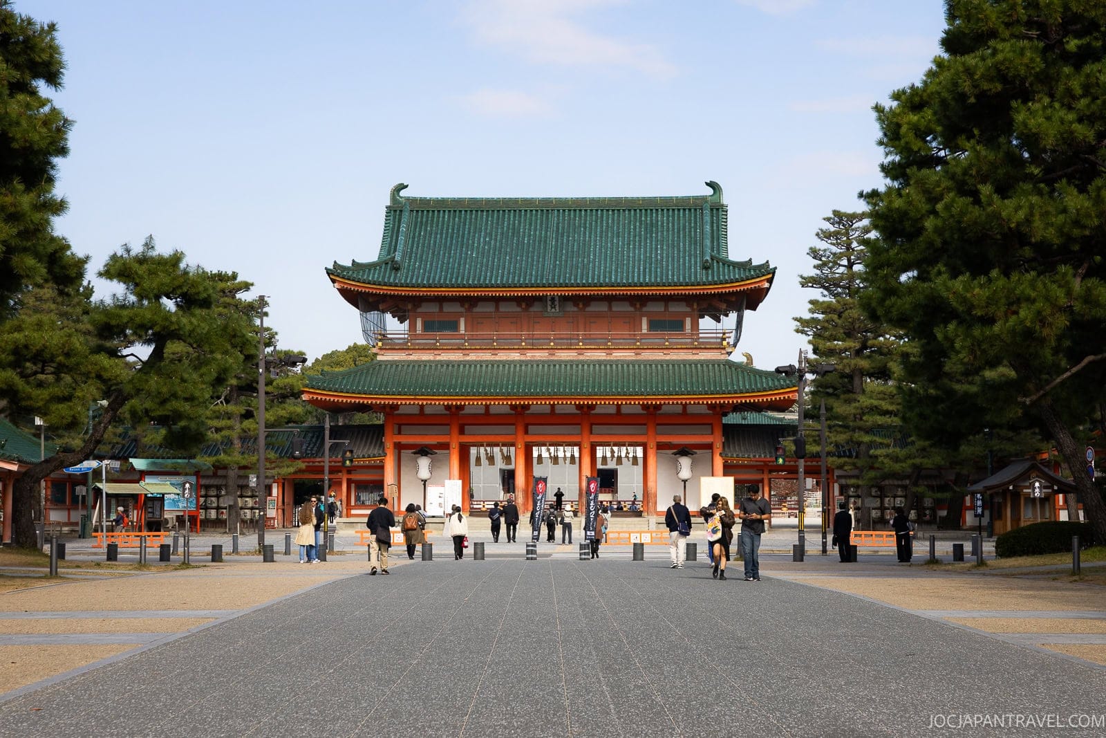 People walk toward the large, historic Heian Shrine gate with its green roof tiles and striking orange pillars, surrounded by trees under a clear Kyoto sky.
