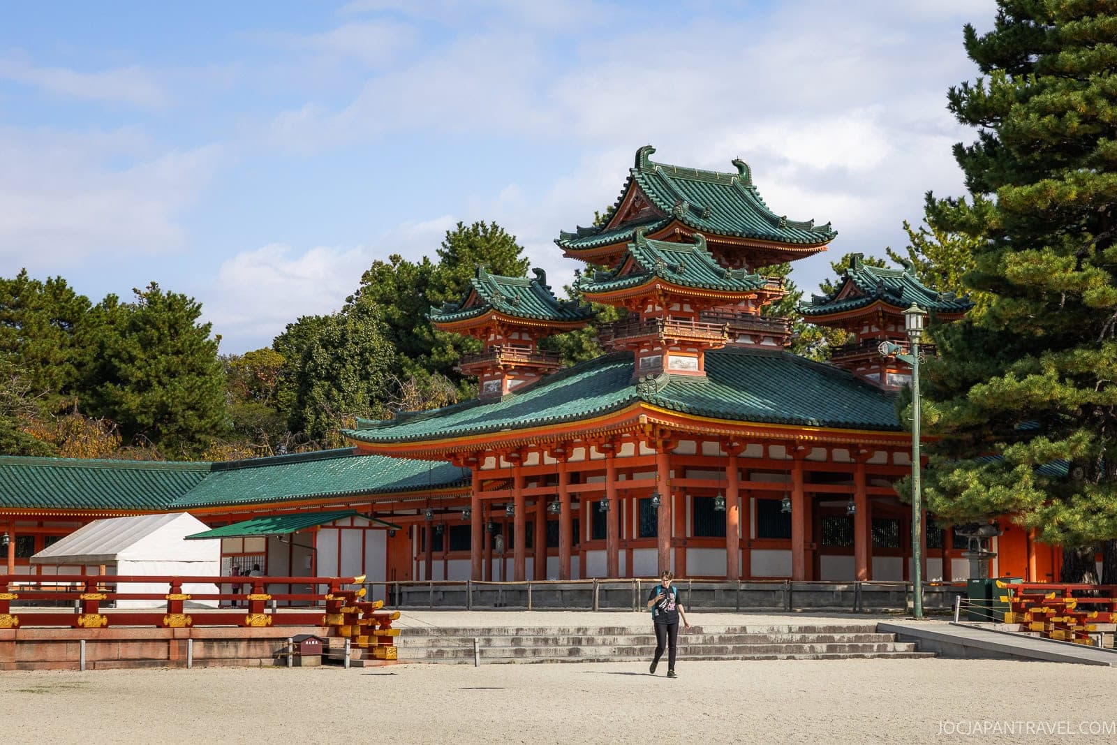 A person stands in front of the historic Heian Shrine in Kyoto, with its striking red columns, green-tiled roofs, and ornate architectural details, surrounded by trees and a sandy courtyard.