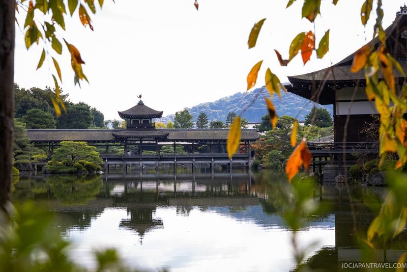 A serene Japanese garden with a traditional wooden pavilion, calm reflective pond, and autumn leaves in the foreground evokes the historic beauty of Kyoto. In the distance, green hills rise under a soft sky, reminiscent of the tranquil landscapes surrounding Heian Shrine.
