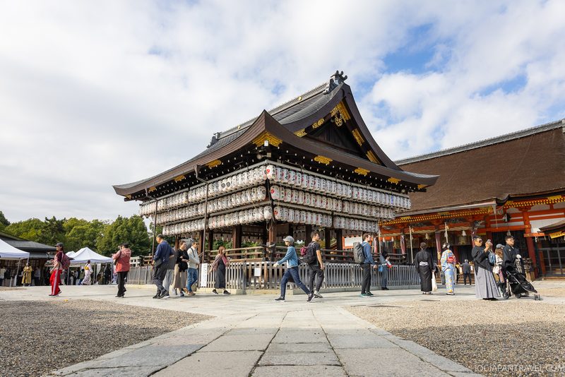 People walk near a traditional Japanese shrine adorned with rows of white paper lanterns under a partly cloudy sky in the heart of Kyoto’s Higashiyama District. Some visitors wear kimonos, while others opt for casual clothing. Trees and stalls are visible in the background.