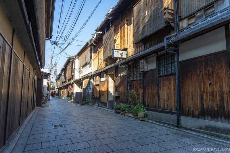 A quiet, narrow street in Kyoto’s Higashiyama District lined with traditional wooden buildings, plants by the doors, and overhead power lines on a clear, sunny day.