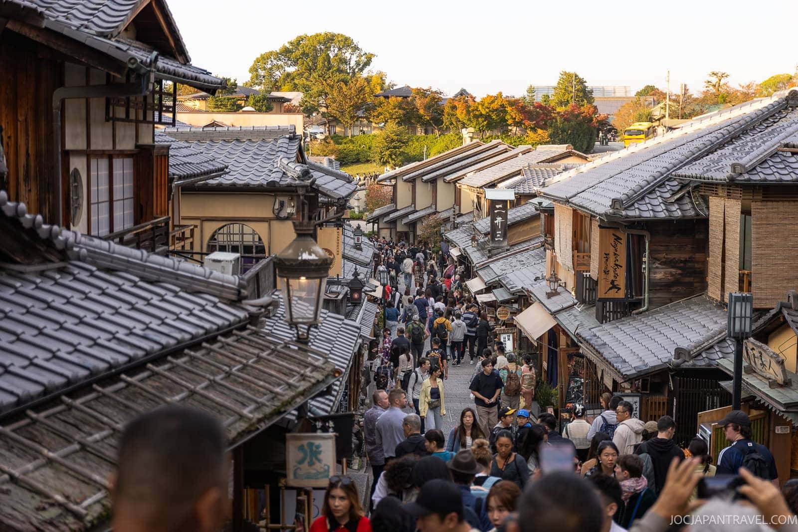 A busy street in Kyoto’s traditional Higashiyama District, lined with wooden buildings and tiled roofs, crowded with people walking and sightseeing on a sunny day.