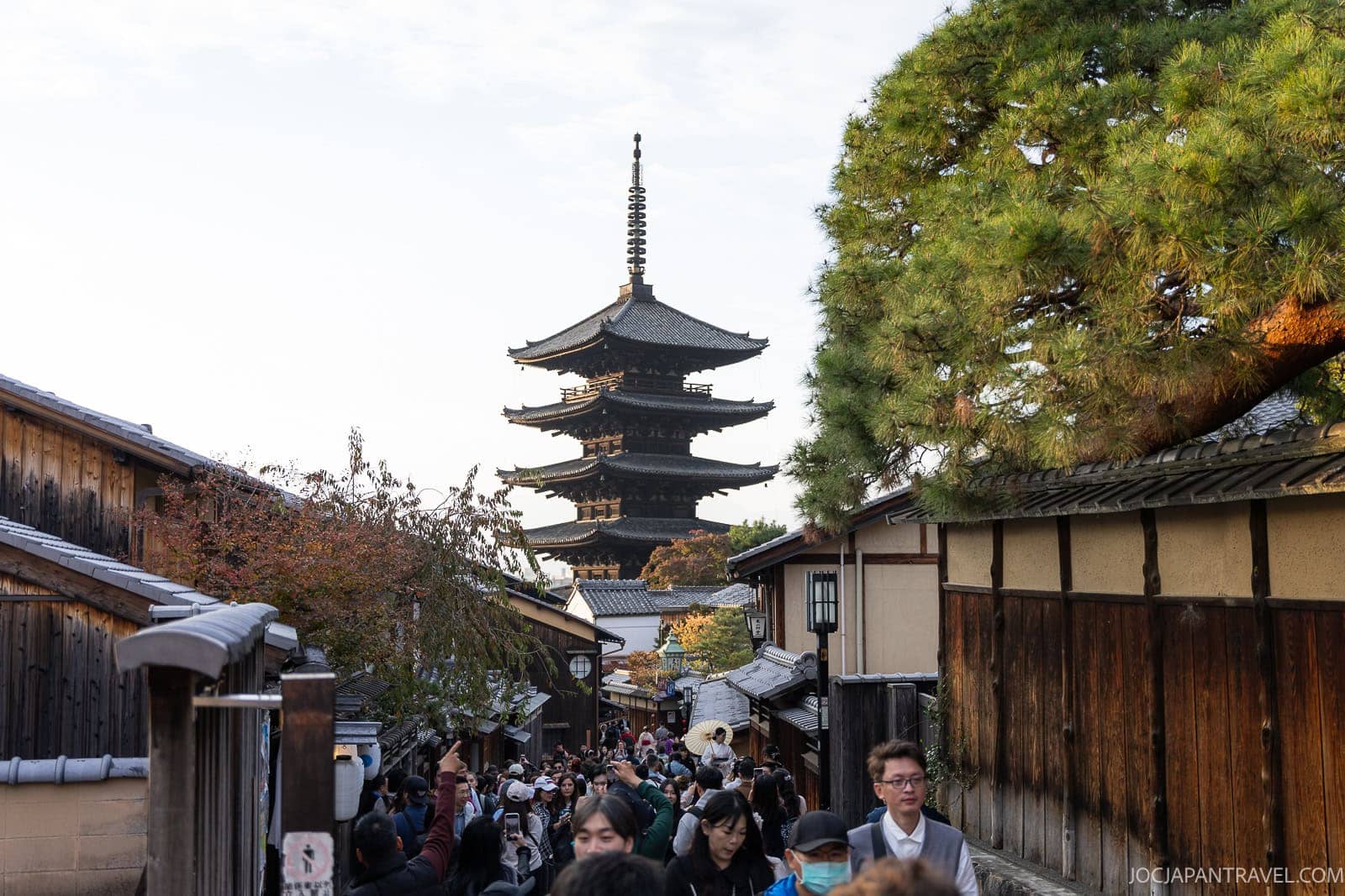 Crowds walk along a narrow street in Kyoto’s historic Higashiyama District, lined with traditional wooden buildings and leading towards a tall, multi-tiered pagoda surrounded by trees in the background.