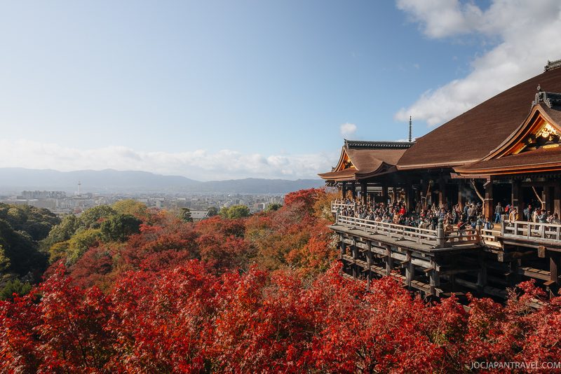 Kiyomizu-dera temple in Kyoto, nestled in the historic Higashiyama District, overlooks a cityscape and distant mountains, surrounded by vibrant autumn foliage and a crowd of visitors enjoying sightseeing on its wooden terrace.