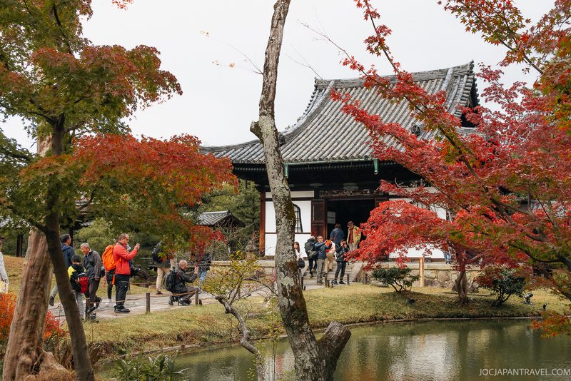 A group of people stand by a pond and colorful autumn trees in front of a traditional Japanese building with a tiled roof, capturing the serene beauty of Kyoto’s Higashiyama District—a renowned tourist destination famed for its historic charm.