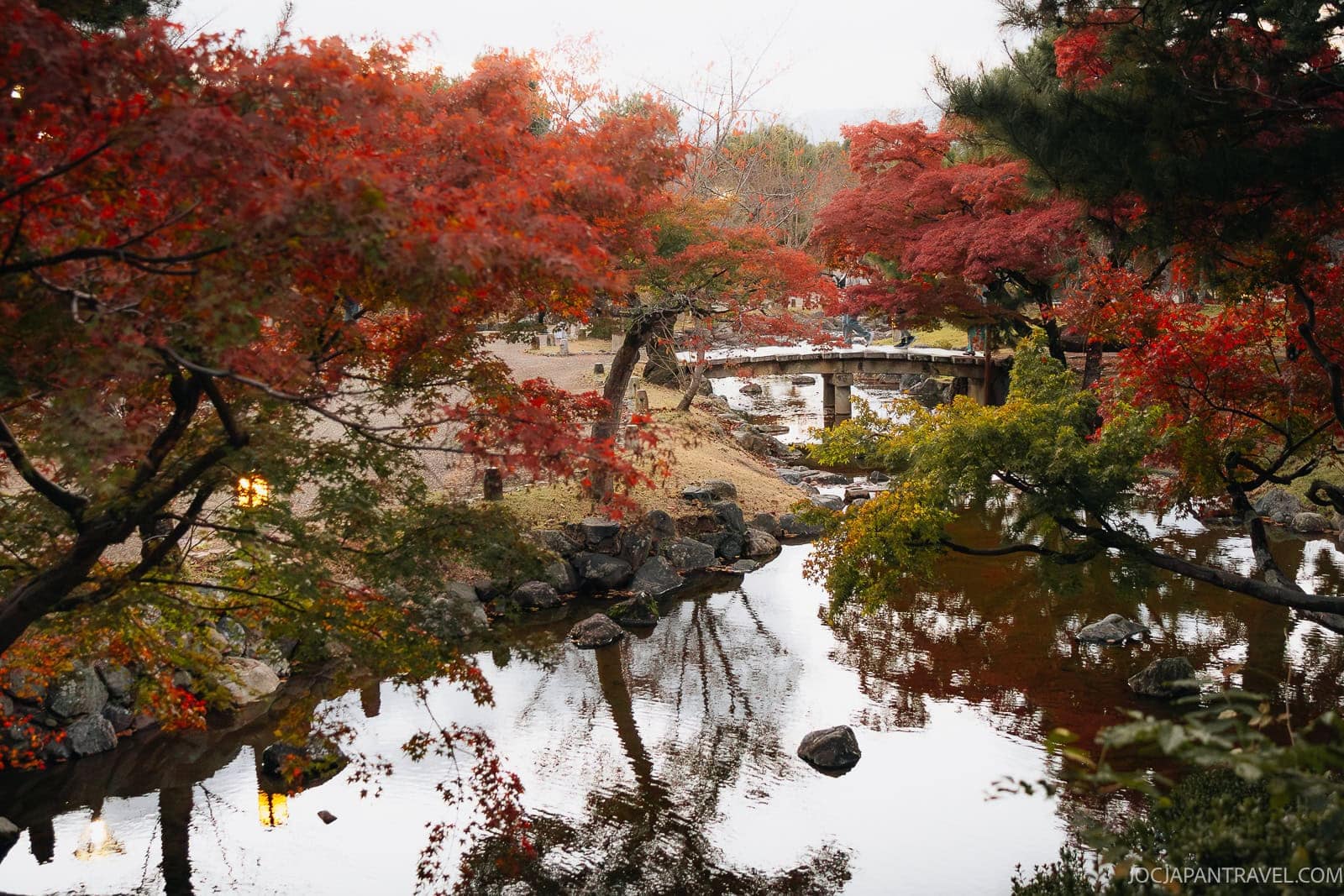 A serene Japanese garden in autumn, nestled in the heart of Kyoto’s historic Higashiyama District, with vibrant red and orange foliage, a calm pond reflecting trees, rocks, and a curved wooden bridge in the background.