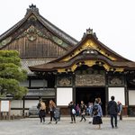 Nijo Castle - Exploring the Palace of the Tokugawa Shoguns Groups of people walk in front of an ornate traditional Japanese building with gold accents, wooden carvings, and a tiled roof, surrounded by trees and gravel paths.