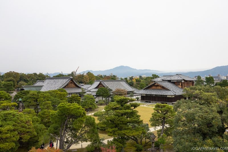 Traditional Japanese buildings with gray tiled roofs are surrounded by lush green trees and garden paths, with mountains visible in the background under a cloudy sky.