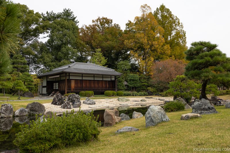A traditional Japanese building stands beside a tranquil garden with rocks, gravel, a pond, and green trees, some with autumn leaves, under a cloudy sky.