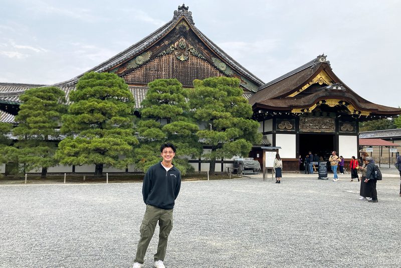 A person stands smiling in front of a historic Japanese building with ornate rooftops and large green trees; several other visitors are nearby under a cloudy sky.