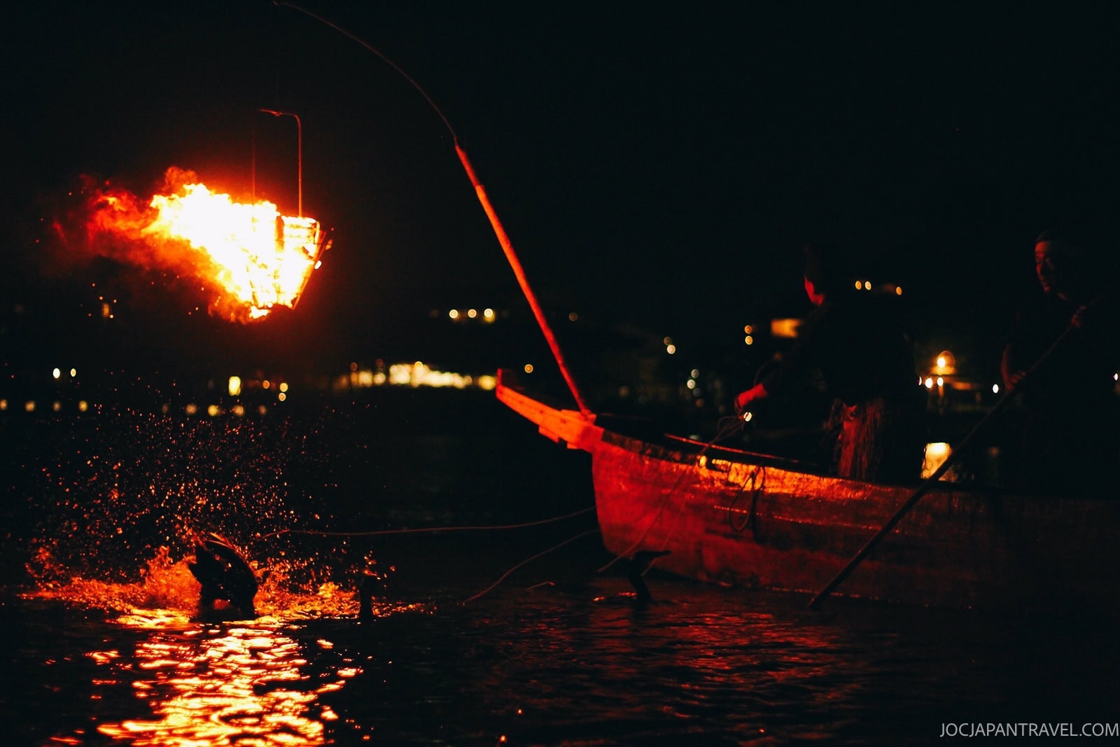 A wooden boat glides along the Uji River at night, with a large flaming torch hanging above the water, illuminating a figure leaning forward and another person from Uji standing with an oar.