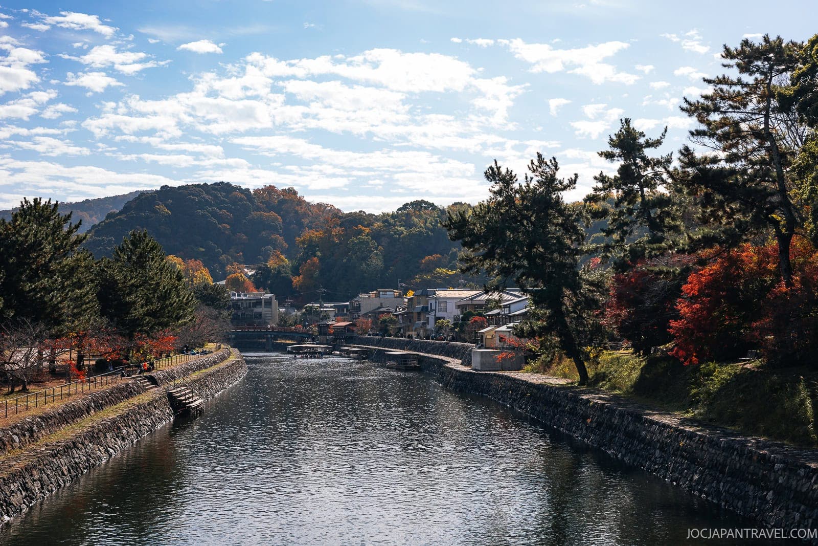 A calm Uji River flows between stone banks lined with trees showing autumn colors, with houses and hills in the background under a partly cloudy sky.