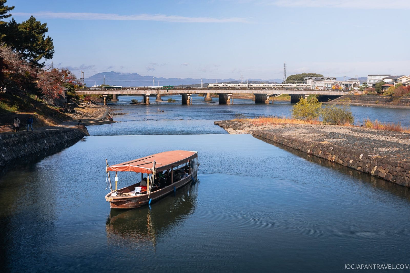 A wooden boat with a canopy floats on the calm Uji River, heading toward a bridge in the distance. Trees and buildings line the riverbanks of Uji under a clear blue sky.
