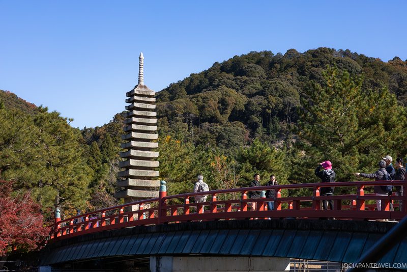 People cross a red bridge over the tranquil Uji River, with a stone pagoda and lush green trees in the background, under a clear blue sky.