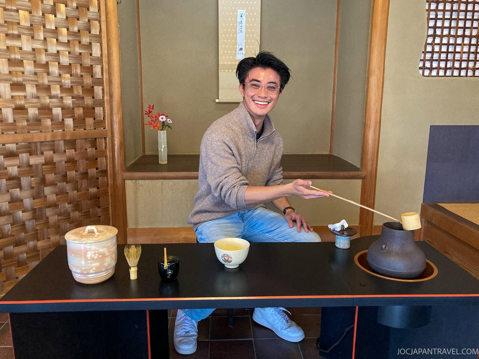 A smiling person sits at a traditional Japanese tea ceremony table, holding a bamboo ladle above a teapot filled with fragrant Uji Tea. Green tea utensils and bowls are arranged neatly in front of them in a cozy, wooden room.