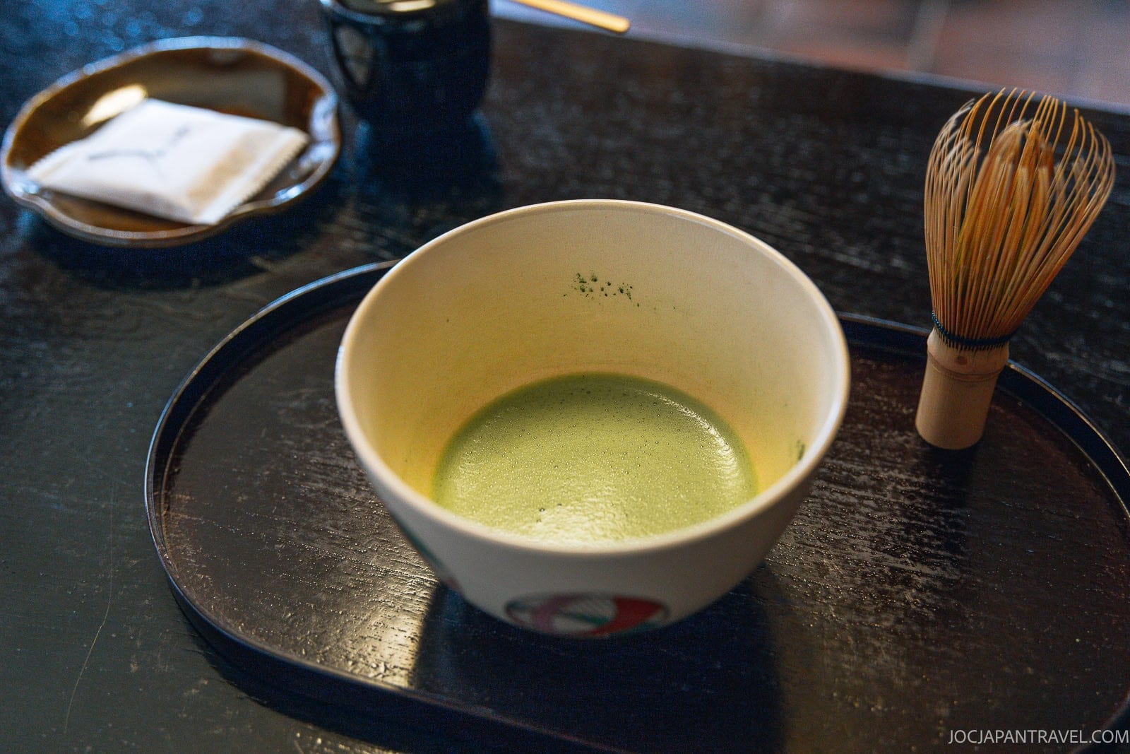 A bowl of matcha tea—celebrated as the home of green tea—sits on a tray with a bamboo whisk and a plate holding a folded napkin, all set on a dark wooden table.
