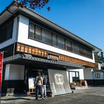 Uji Tea - The Home of Green Tea People stand outside a traditional Japanese building with white walls, wooden accents, and a large black noren curtain at the entrance. A red banner and autumn leaves are visible, suggesting a restaurant or shop—perhaps even the home of green tea, where visitors can enjoy authentic Uji Tea in an inviting setting.