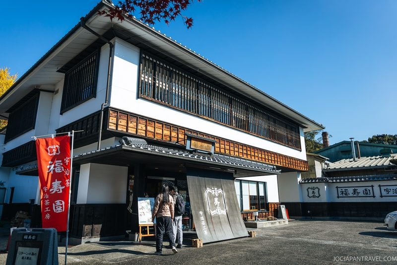 People stand outside a traditional Japanese building with white walls, wooden accents, and a large black noren curtain at the entrance. A red banner and autumn leaves are visible, suggesting a restaurant or shop&mdash;perhaps even the home of green tea, where visitors can enjoy authentic Uji Tea in an inviting setting.